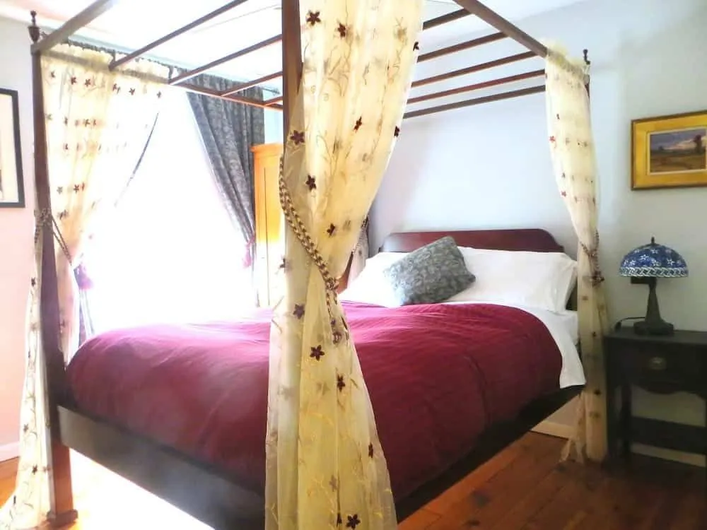 Queen canopy bed in the Carmen Suite with red bedding, historic wood floors, and natural light from a window