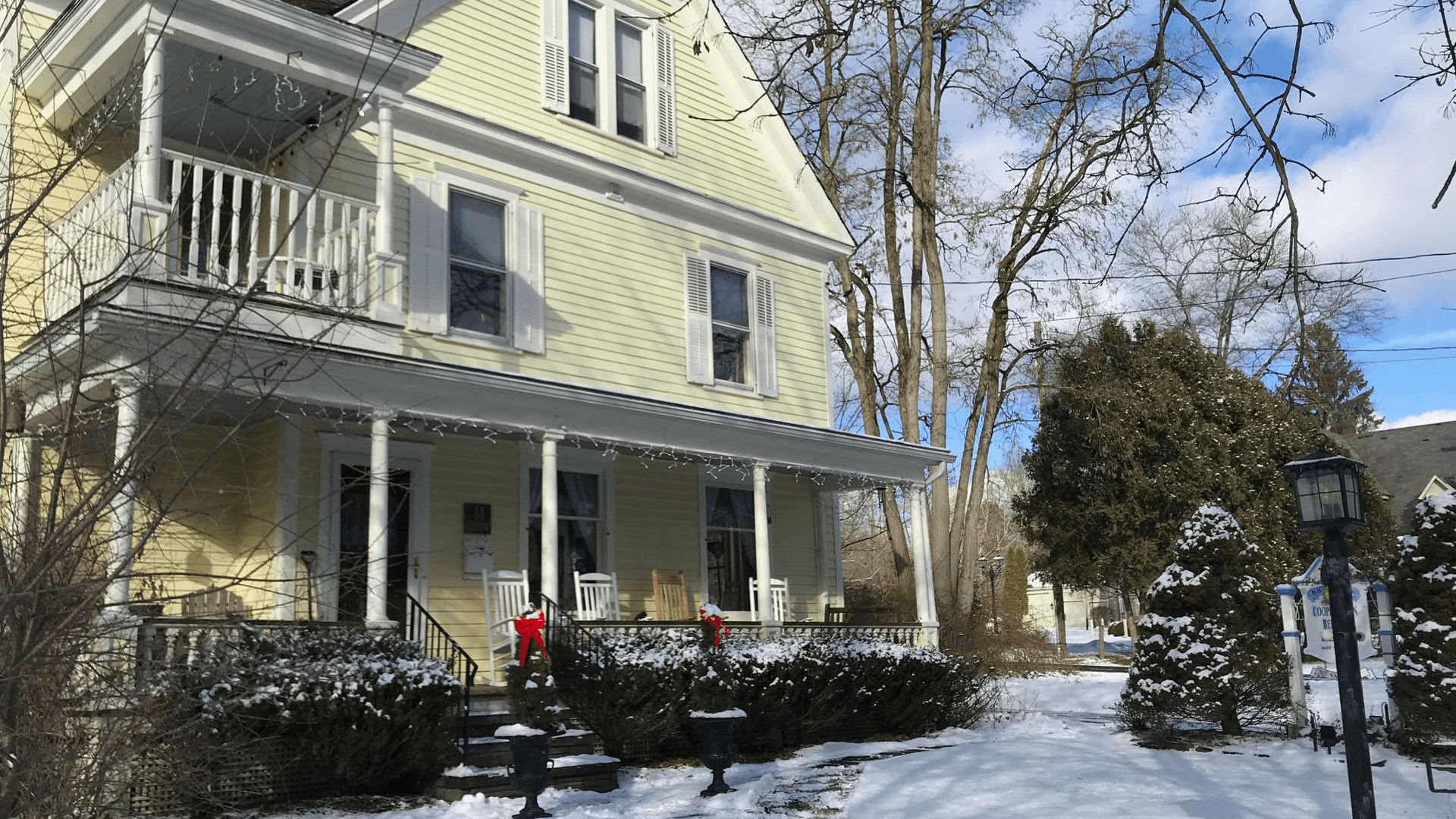 A yellow two-story house is surrounded by snow and trees on a sunny winter day.