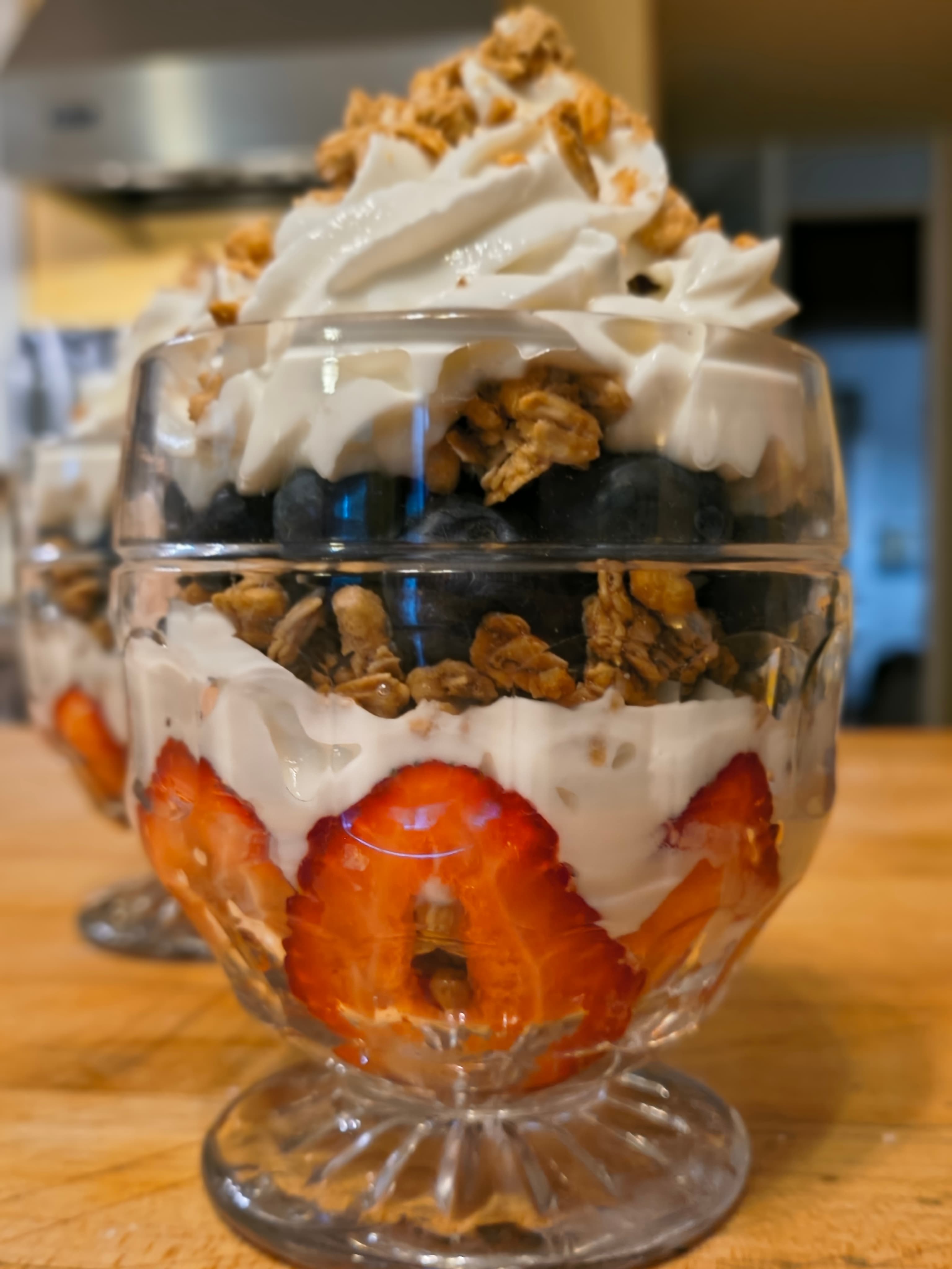 A clear glass dessert cup layered with strawberries, granola, blueberries, and whipped cream.