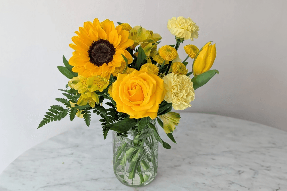 A bright bouquet of yellow flowers, including sunflowers, roses, and tulips, arranged in a clear glass vase.