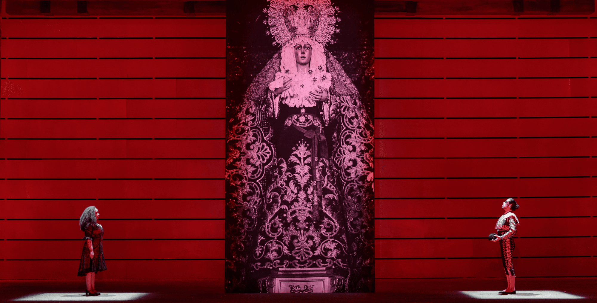 A dramatic scene featuring two figures in elaborate costumes standing before a large, ornate backdrop in a red-themed setting.