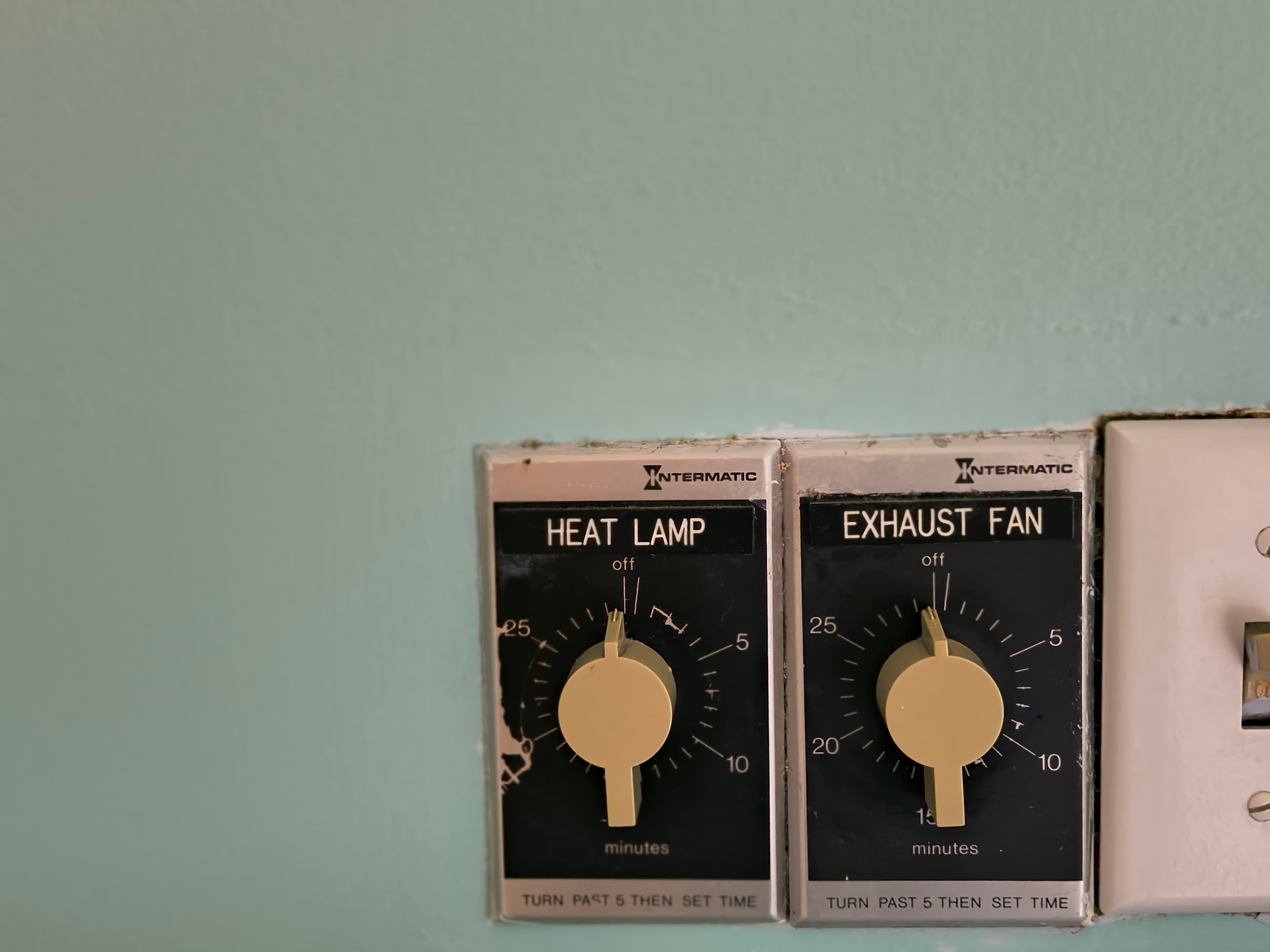 Two timer controls for a heat lamp and exhaust fan mounted on a light green wall.