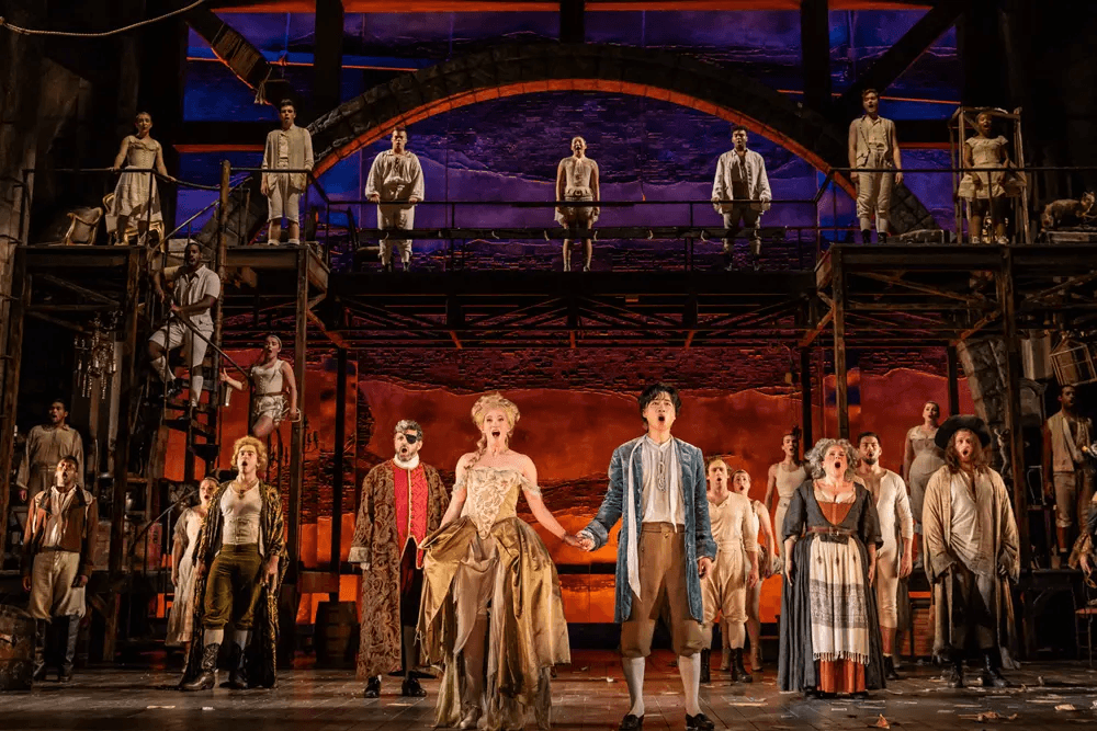 A diverse cast of actors performing on stage, with a dramatic backdrop and colorful lighting.