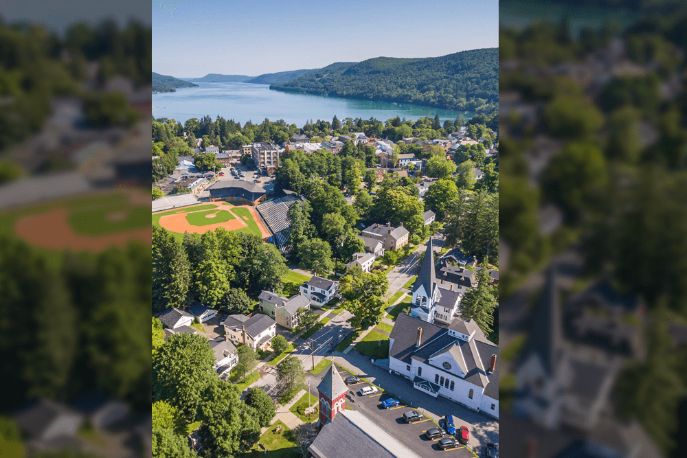Aerial view of a town with a baseball field, houses, and a lake surrounded by hills.
