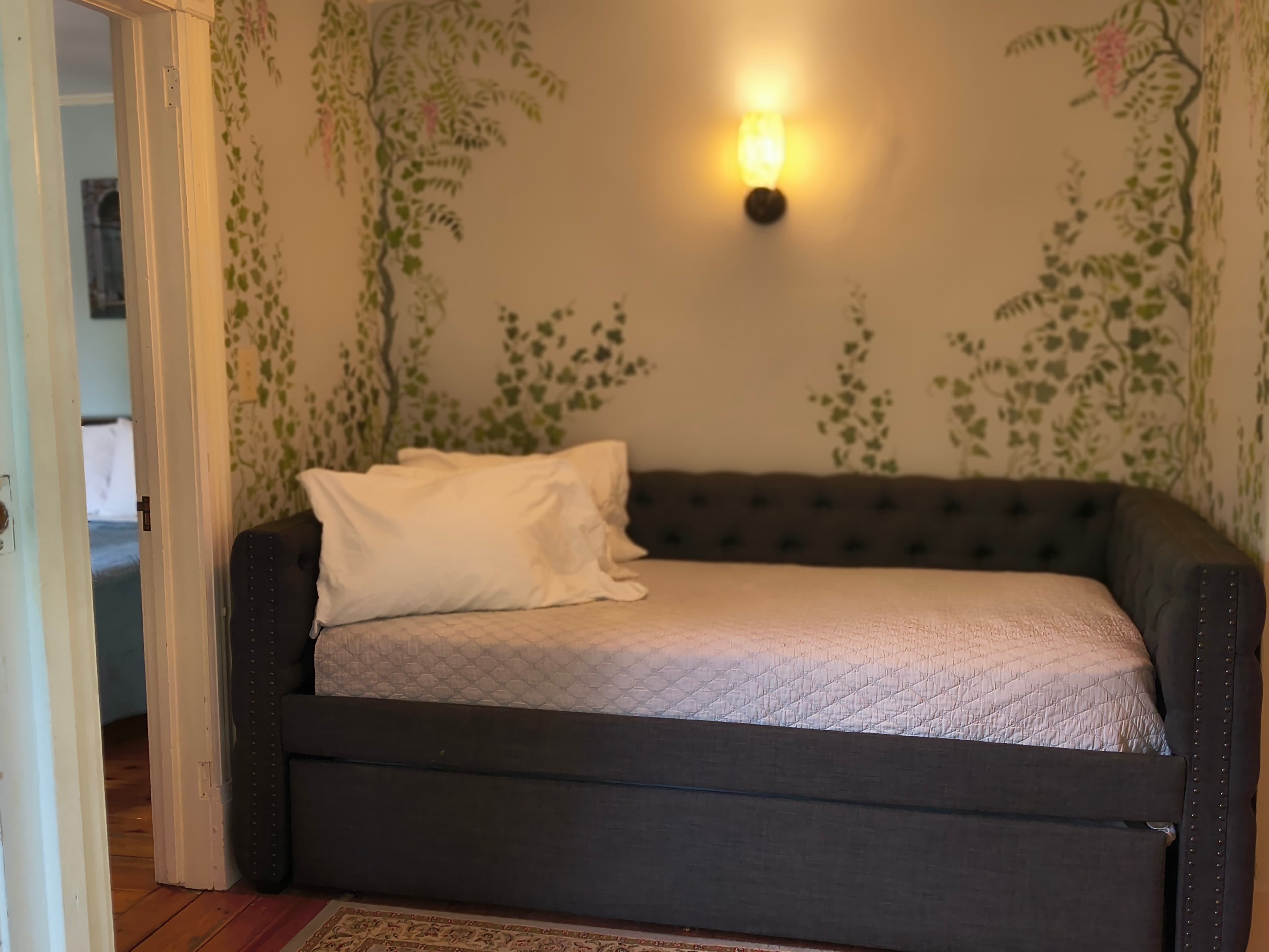 A cozy room with a dark grey daybed, decorative pillows, and leafy mural walls.