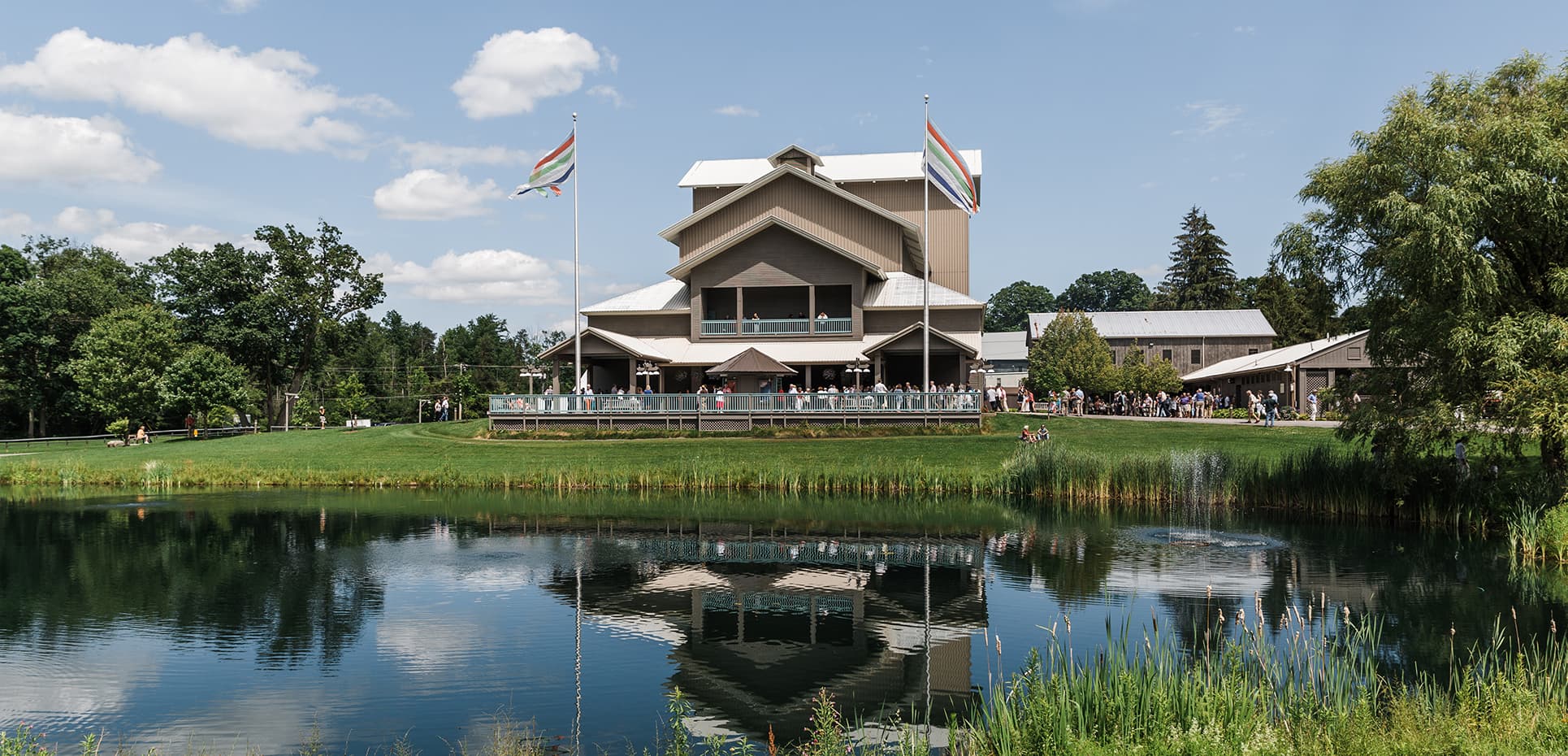 A modern building with flags reflected in a pond, surrounded by greenery and blue sky.