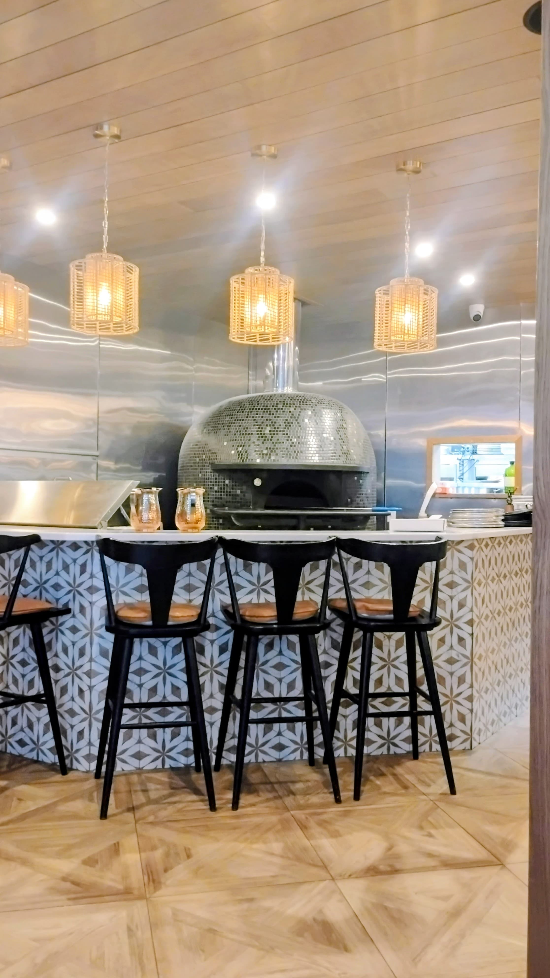 A modern bar area with black stools, patterned tiles, and a stone pizza oven under warm pendant lighting.