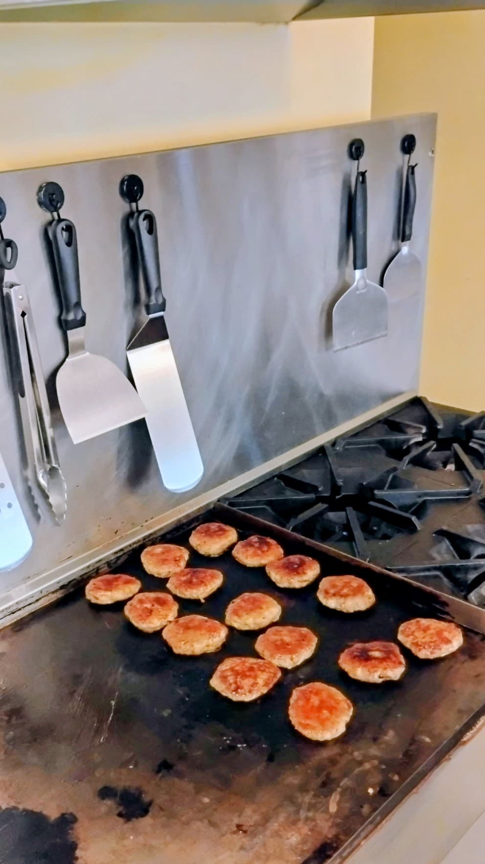 A griddle with golden-brown patties and cooking utensils hanging on a stainless steel backsplash.