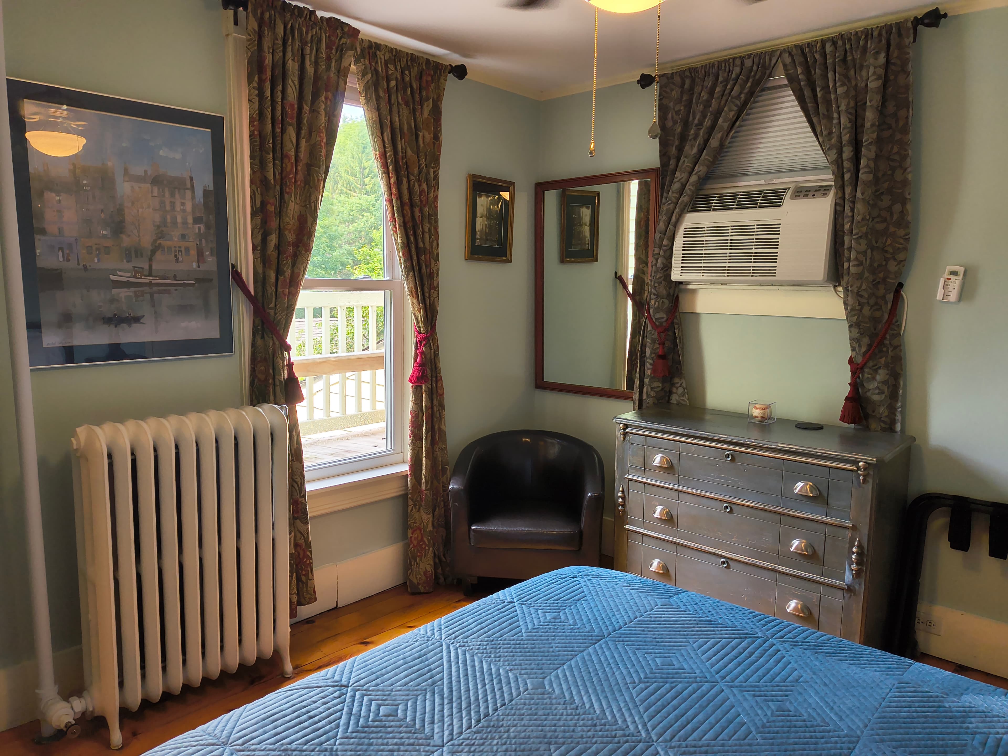 Cozy bedroom featuring a blue quilt, dresser, chair, and large window with floral curtains.