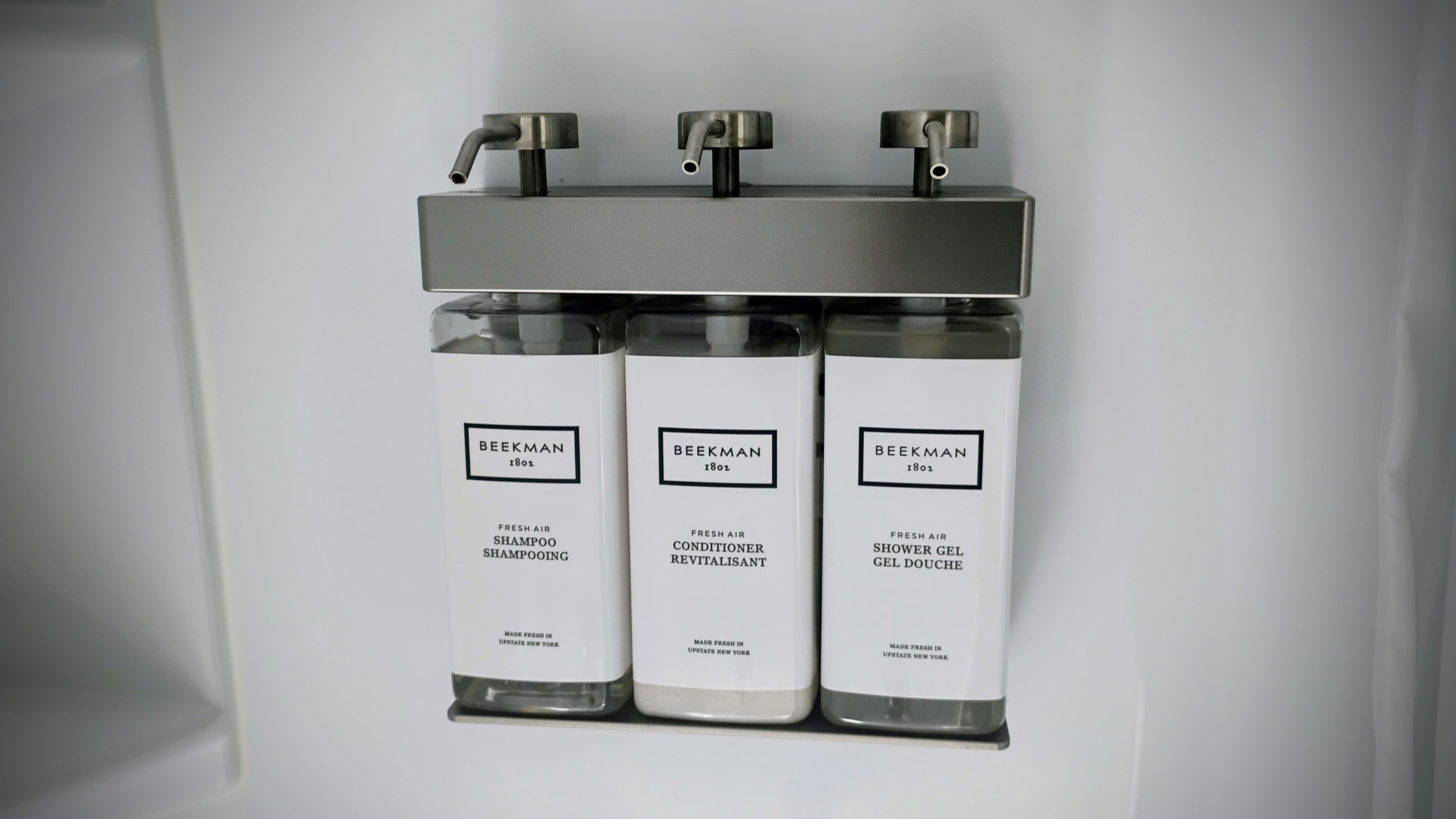 Dispenser with three clear bottles labeled shampoo, conditioner, and shower gel.