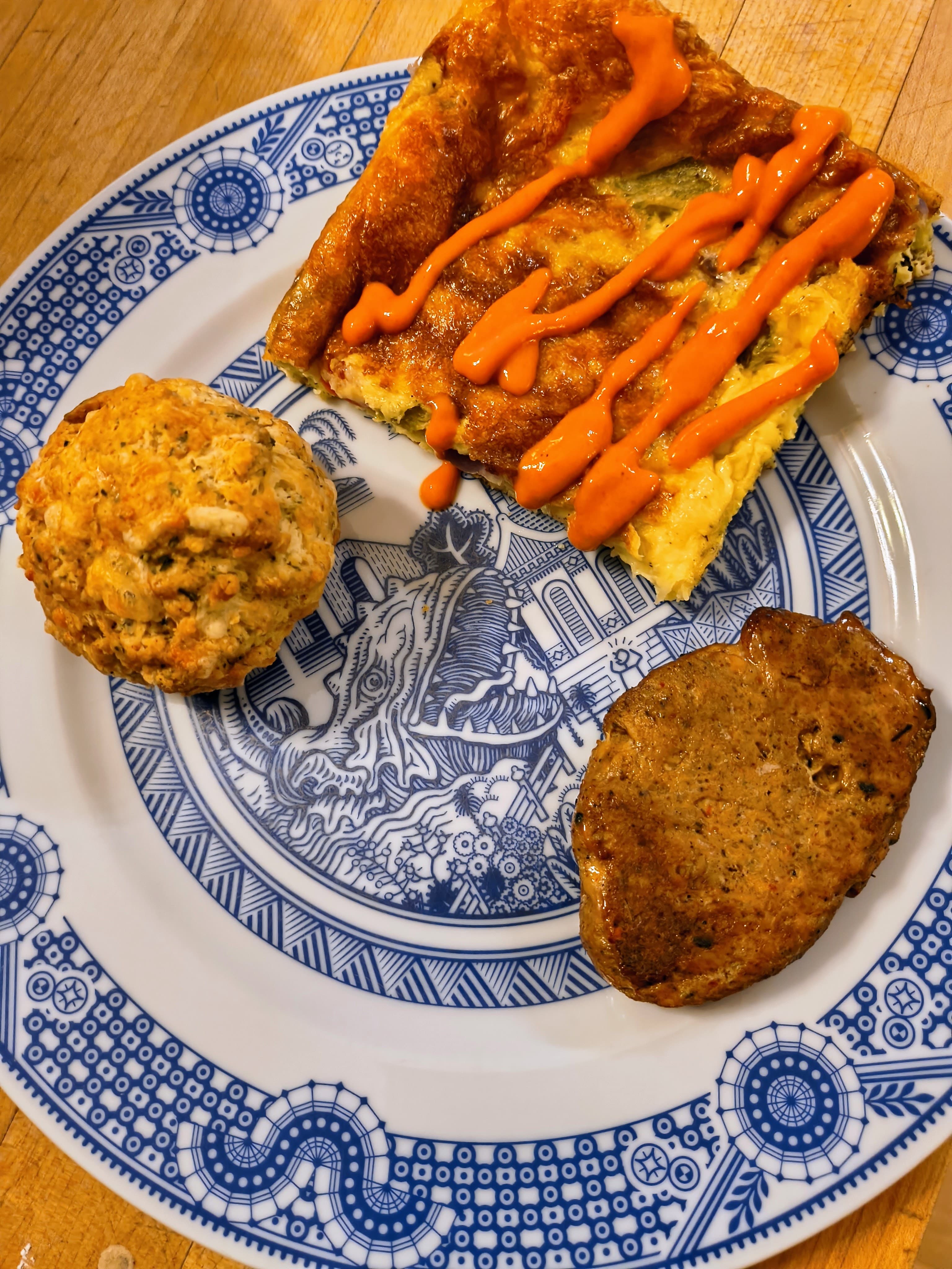 A plate with a slice of frittata drizzled with sauce, a muffin, and a meat patty.