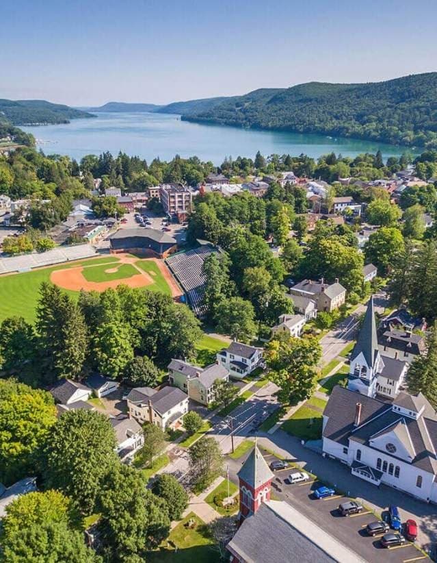 Aerial view of a small town with baseball field, houses, and a lake surrounded by green hills.