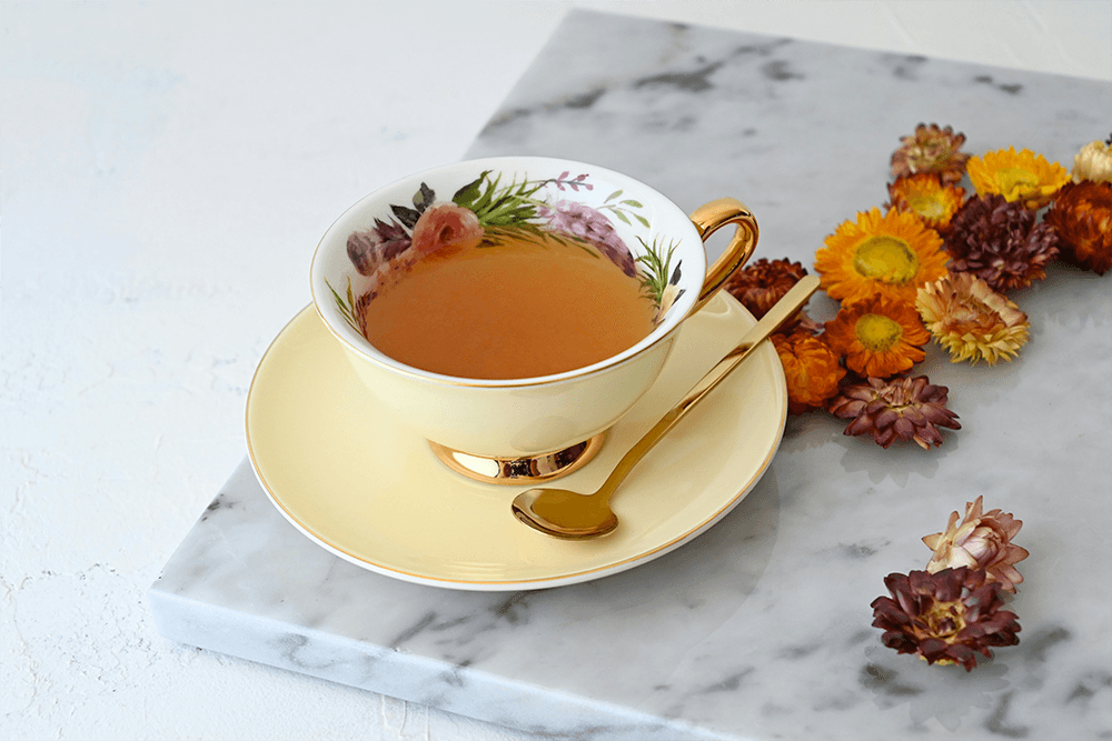 A decorated teacup with tea sits on a saucer beside dried flowers on a marble surface.