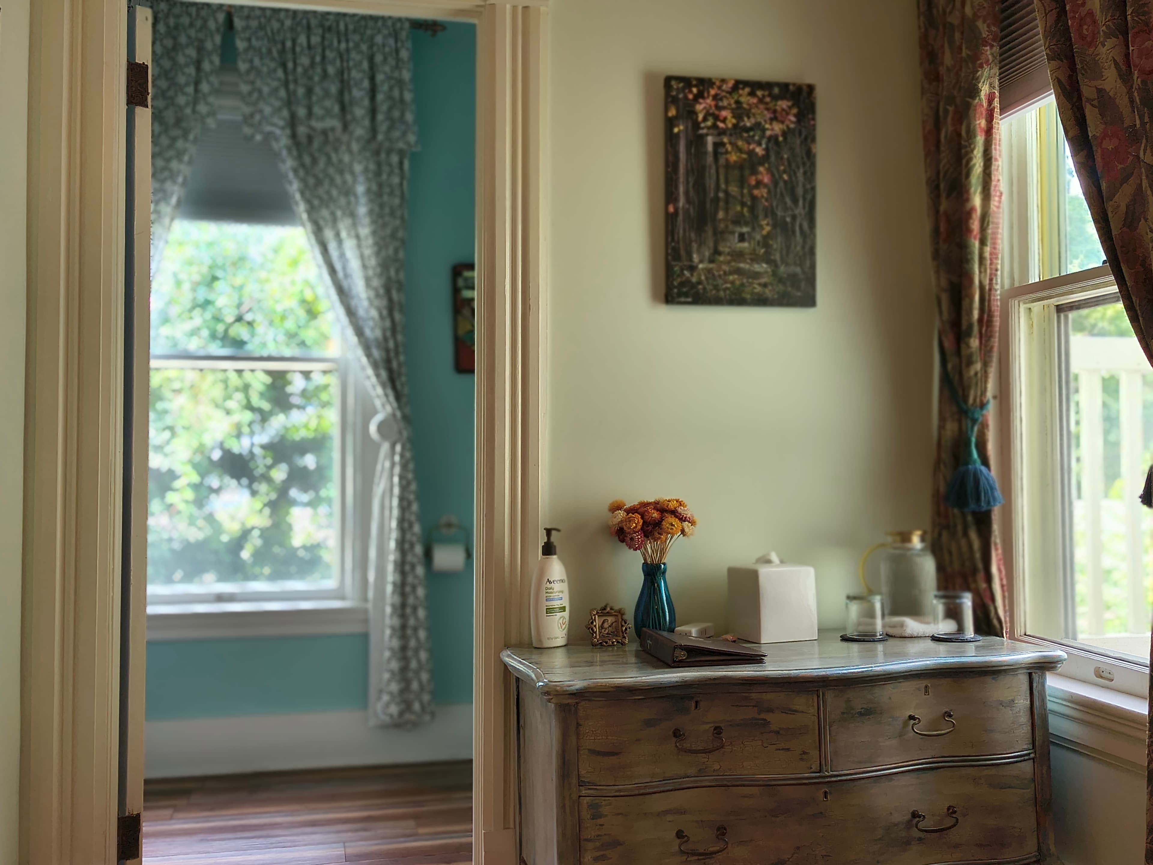 A cozy room with a wooden dresser, vase of flowers, and a view of a bright window through a doorway.