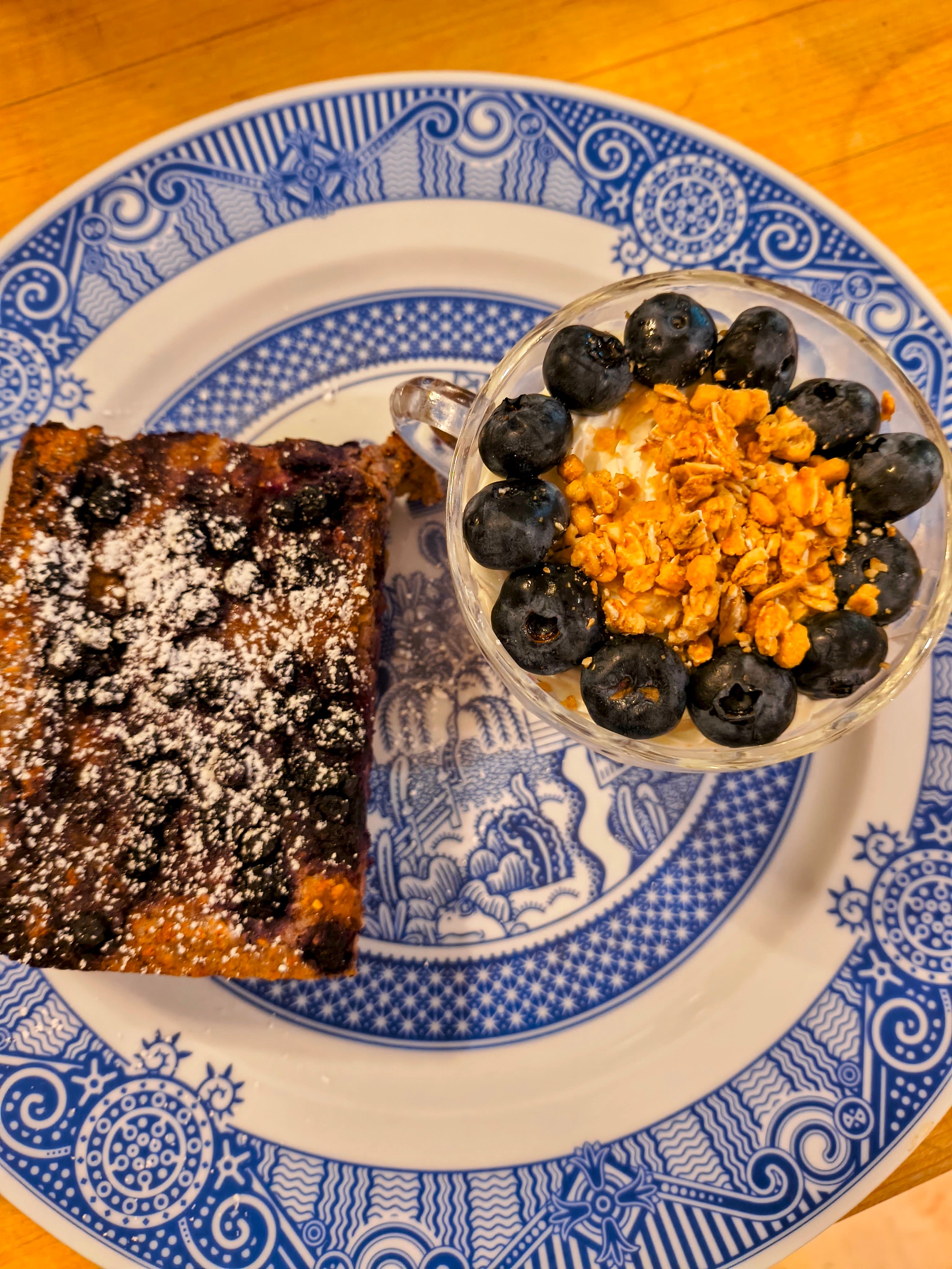 A slice of blueberry dessert sits next to a glass of yogurt topped with blueberries and granola on a blue-patterned plate.