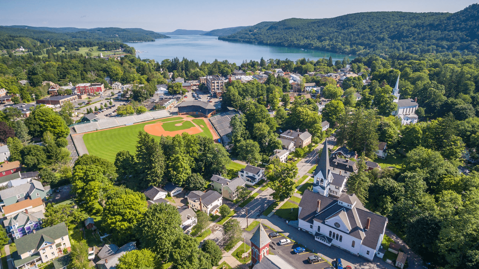 Aerial view of a small town with a baseball field, surrounded by lush greenery and a serene lake.