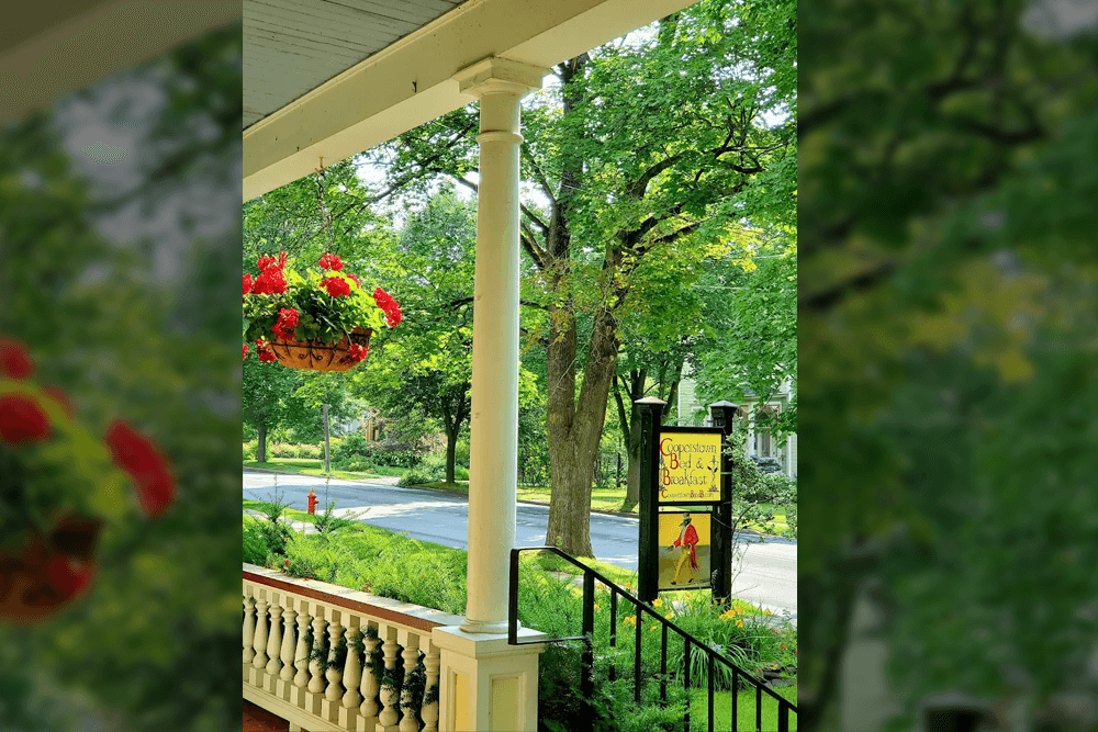 A porch view featuring a hanging flower basket, a white column, and a sign for a bed and breakfast surrounded by greenery.